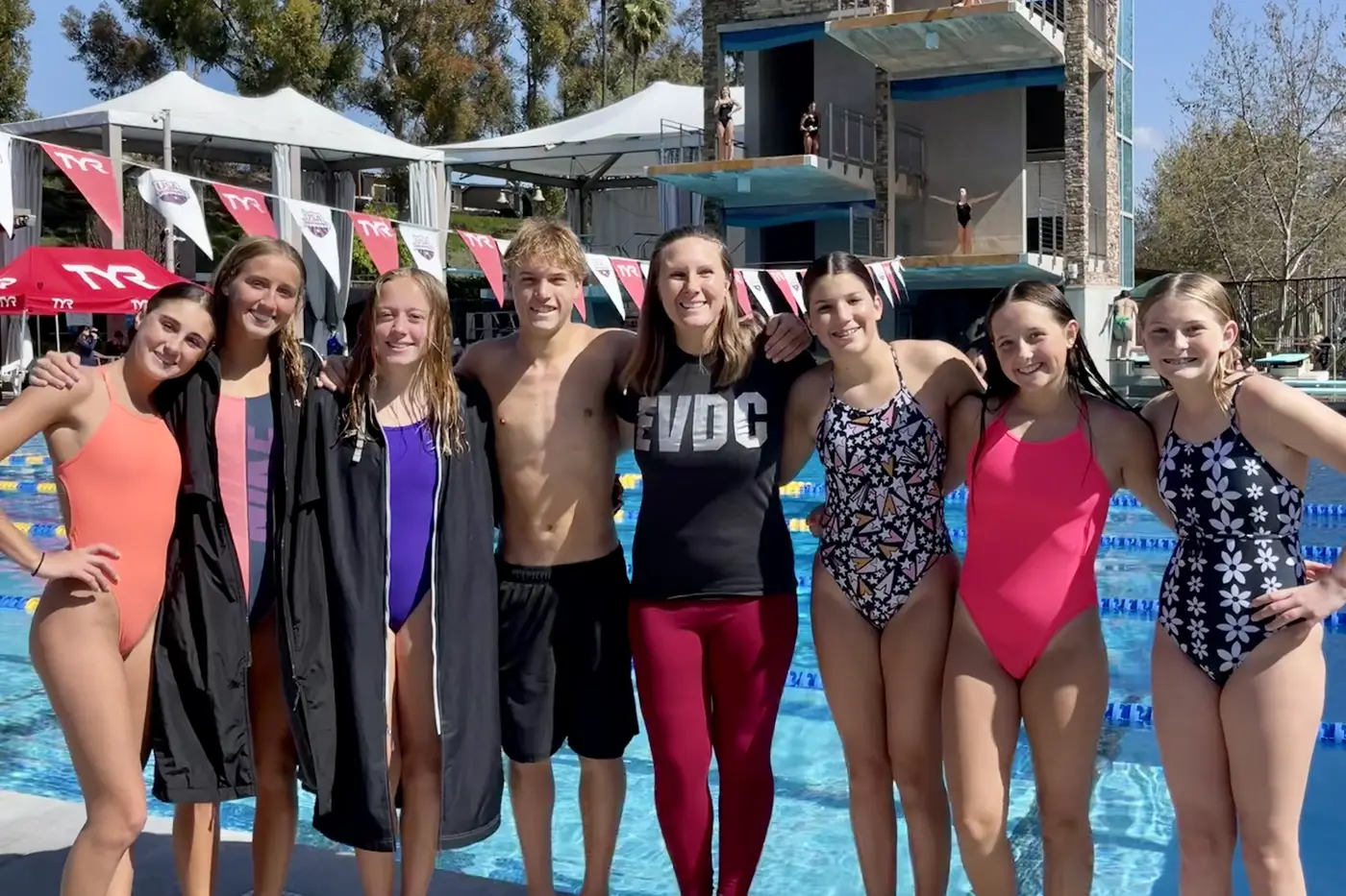 The team and a coach on the deck at a meet, banners strung behind them and a diving tower beyond.