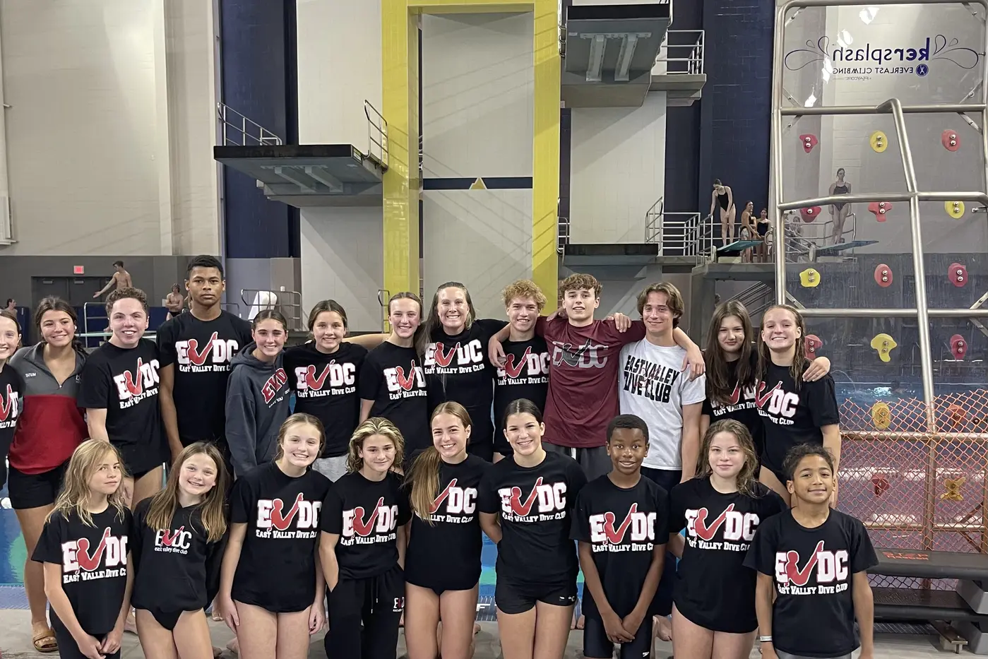 The club in team shirts at an indoor aquatic center, diving platforms behind them.