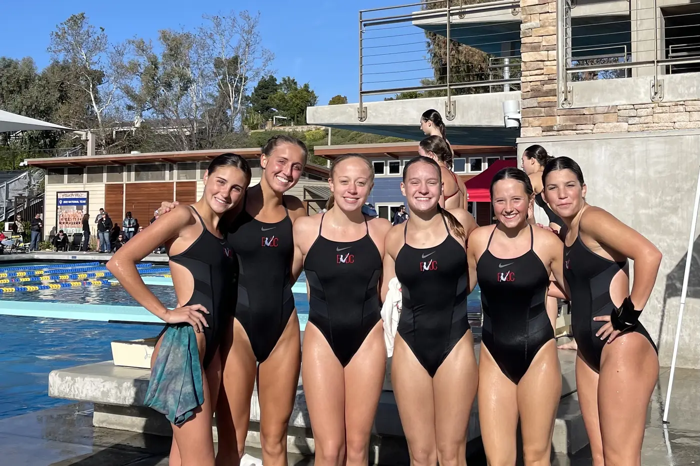 Divers in team suits on the deck at an outdoor meet, a diving tower rising behind.