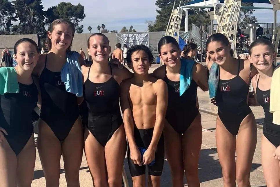 Divers in team suits with towels over their shoulders, poolside at an outdoor meet.